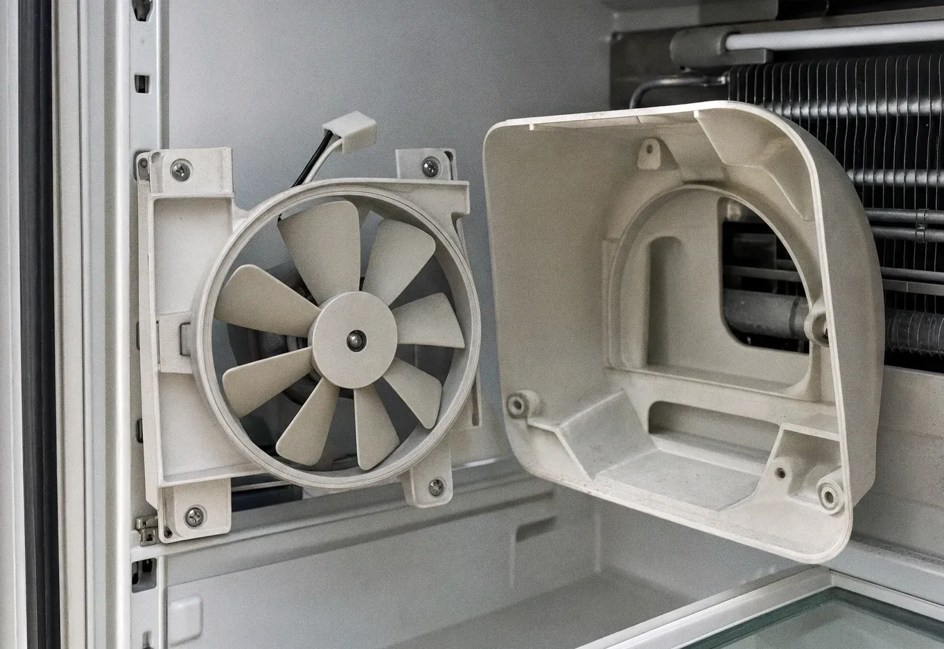 A circulation fan behind its opened cover inside an appliance compartment, with a finned coil alongside it