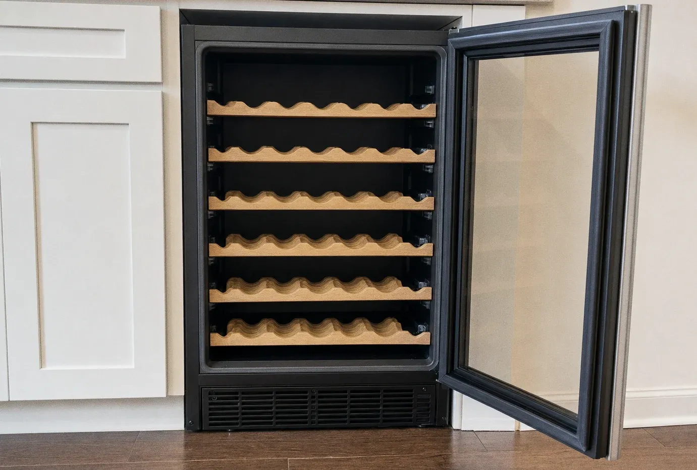 A free-standing wine cabinet with empty wooden racks and its glass door swung wide open beside pale cabinetry