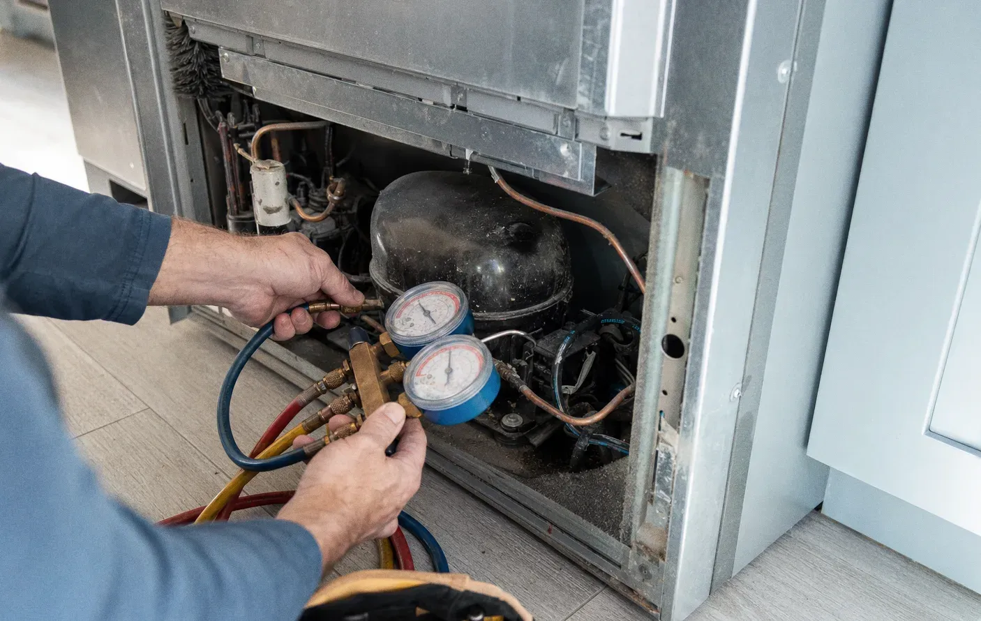 A hand on manifold gauge valves at the back of an opened cabinet, compressor and copper pipework behind