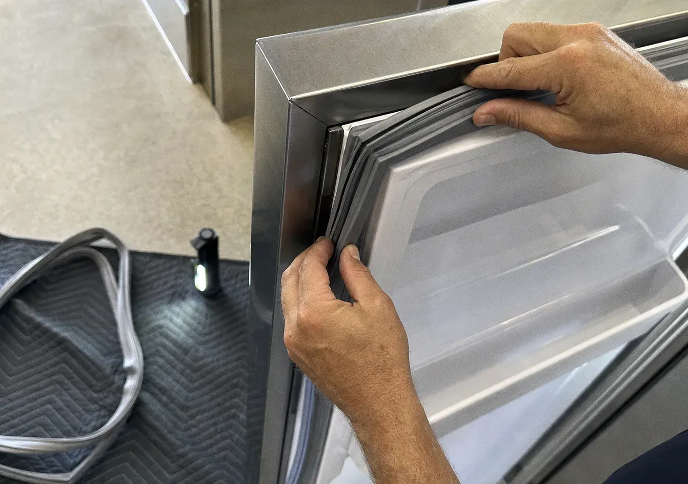 Hands lifting a magnetic gasket away from the edge of a stainless door, a coil of tubing on the mat below